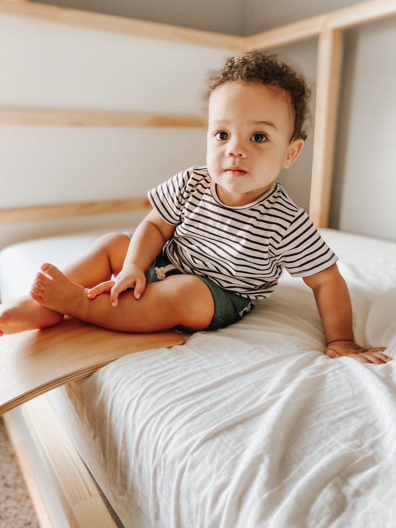 May include: A young child with brown curly hair sits on a white bed wearing a black and white striped shirt and blue denim shorts. The child is sitting on a wooden balance board.