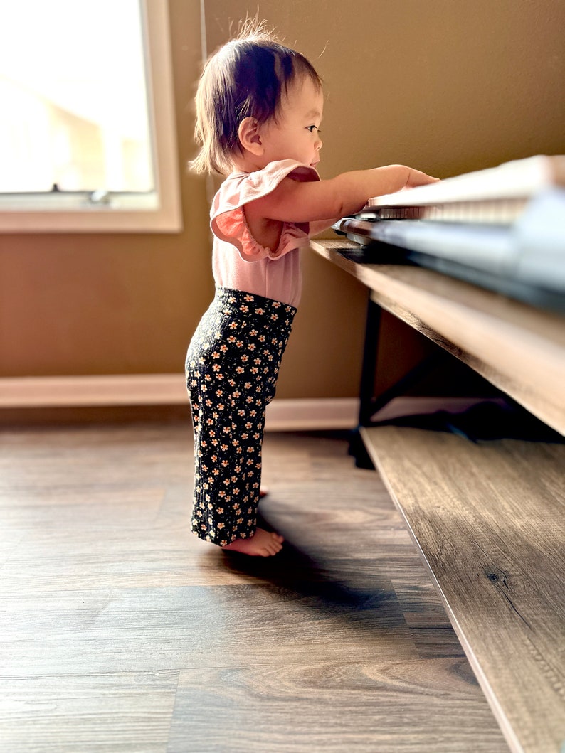 May include: A young child wearing a pink ruffled shirt and black floral pants stands on a wooden floor and reaches for a keyboard on a wooden table.