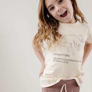 May include: A young girl wearing a light beige t-shirt with a black line drawing of a dinosaur and the word "par-a-saur-ol-o-phus" printed on it. The shirt also has the definition of the word printed below the dinosaur. The girl is wearing brown pants with a white drawstring.