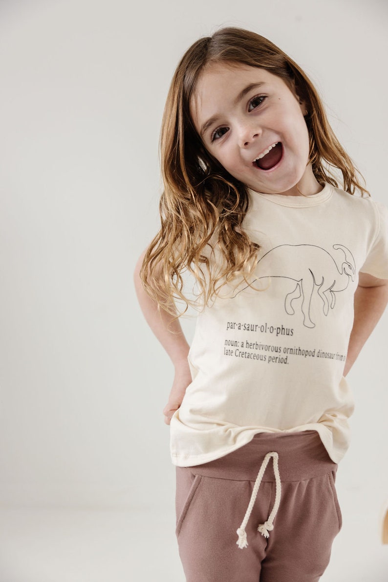 May include: A young girl wearing a light beige t-shirt with a black line drawing of a dinosaur and the word "par-a-saur-ol-o-phus" printed on it. The shirt also has the definition of the word printed below the dinosaur. The girl is wearing brown pants with a white drawstring.