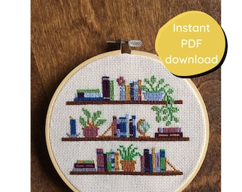 May include: Cross-stitch embroidery of a bookshelf design with books and plants. The design is in a wooden embroidery hoop. The text "Instant PDF download" is in a yellow circle. The background is a wooden surface.