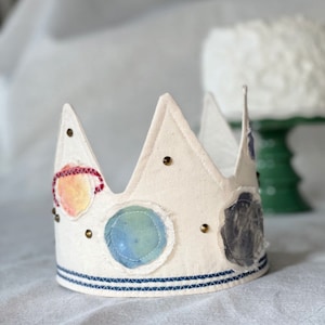 May include: A white felt crown with a blue and white striped band. The crown is decorated with colorful fabric circles and gold studs.