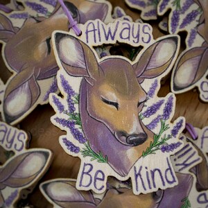 May include: Wooden keychain with a purple and brown deer illustration. The keychain has the text "Always Be Kind" around the deer.