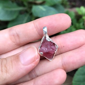以下が含まれることがあります： 銀製のベゼルにセットされた、生のピンク色のクリスタルペンダント。クリスタルは、粗く自然な形をしており、濃いピンク色をしています。