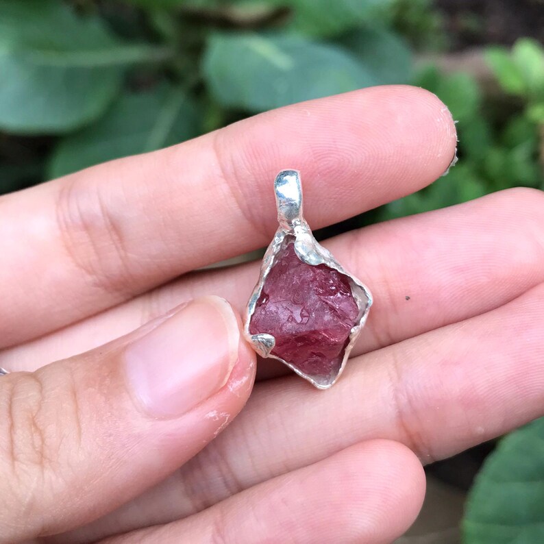 以下が含まれることがあります： 銀製のベゼルにセットされた、生のピンク色のクリスタルペンダント。クリスタルは、粗く自然な形をしており、濃いピンク色をしています。