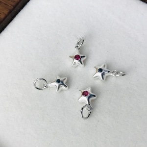 May include: Four small silver star-shaped charms, each featuring a single embedded gemstone. Two charms contain a vibrant red stone, while the other two charms feature a dark blue stone. Each star charm includes a small circular jump ring for attachment.