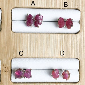 May include: Four pairs of earrings with raw ruby gemstones. The earrings are displayed on a wooden board with white felt lining. The earrings are labeled A, B, C, and D.