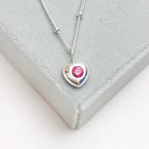 Op de afbeelding: Een zilveren hartvormige hangerketting met een helderroze edelsteen in het midden. De hanger hangt aan een fijne zilveren ketting met kleine kraalaccenten. De ketting wordt gepresenteerd op een grijze ondergrond.