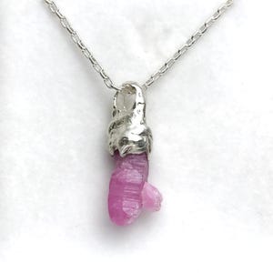 May include: A silver chain necklace featuring a raw, pink crystal pendant. The crystal is a vibrant shade of pink and has a natural, irregular shape. The pendant is attached to a delicate silver chain, creating a simple yet elegant piece of jewellery.