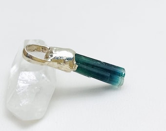 May include: A natural tourmaline pendant with a silver-coloured metal bail. The tourmaline crystal is a deep teal colour with a polished, rectangular shape. The pendant is displayed on a clear, crystalline surface.