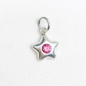 May include: A silver star-shaped charm with a bright pink gemstone in the center. The charm has a small loop at the top for attaching to a necklace or bracelet. The charm is on a white background.