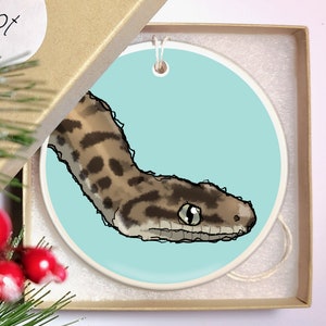 May include: A ceramic Christmas ornament featuring a cartoon illustration of a snake with brown and black markings on a light blue background.