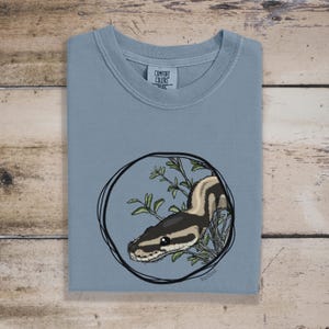 May include: A light blue t-shirt featuring a circular illustration of a snake with a black and tan pattern, surrounded by green leaves. The shirt has a crew neck and the brand "Comfort Colors" is visible on the tag.