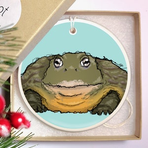 May include: A round ceramic ornament featuring a cartoon illustration of a green frog with large eyes and a wide smile. The frog is on a light blue background.