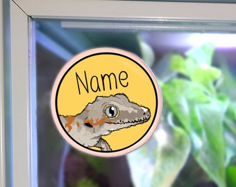 May include: A round sticker with a yellow background and a pink border. The sticker features a cartoon gecko with orange and brown markings and the word "Name" in black text.