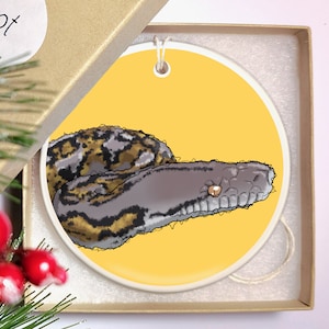 Peut inclure: Une boule de Noël ronde en céramique avec un fond jaune représentant une illustration dessinée à la main d'un serpent python avec des marques noires et brunes. Le serpent est enroulé sur lui-même, la tête tournée vers la droite de l'ornement.