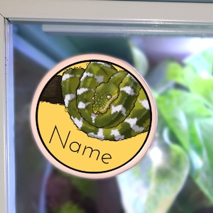 May include: A circular sticker showing a green and white snake on a yellow background. The word "Name" is written in black script. The sticker is set against a backdrop of green plants.