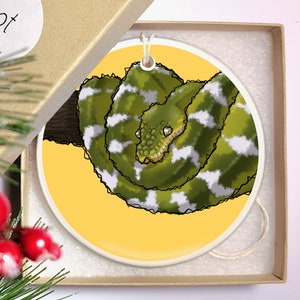 May include: A round ceramic ornament featuring a green and white snake with a yellow background. The snake is curled up on a branch.