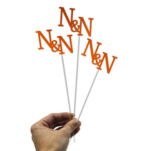 May include: Four orange paper cupcake toppers with the letters "N&N" on each. The toppers are on white sticks and are held in a hand.