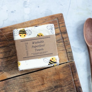 May include: A stack of two white cotton dish towels with a bee pattern. The towels are folded and have a brown paper label that reads "Washable Paper(less) Towels Eco-friendly paper towel replacement handmade in Charolette, NH".