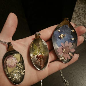 Puede incluir: Tres colgantes con forma de cuchara con flores secas encapsuladas en resina transparente. Los colgantes tienen cuencos de cuchara de metal plateado y mangos de metal dorado. Las flores son rosas, azules y rojas, con detalles de pan de oro. Cada colgante cuelga de una cadena plateada.