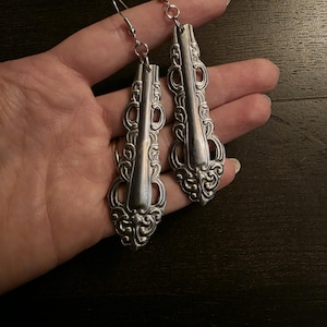 May include: Silver-toned, ornate earrings crafted from repurposed silverware. The earrings feature a decorative, vintage-inspired design with intricate detailing. The hooks are silver and the earrings are held in a hand.