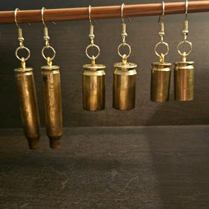 May include: Six pairs of gold bullet casing earrings hanging from a wooden dowel rod. The earrings are made from real bullet casings and have a gold finish.