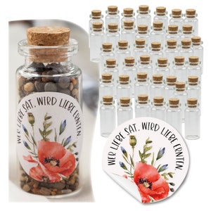 Könnte beinhalten: Eine Sammlung kleiner, klarer Glasflaschen mit Korken. Eine Flasche ist mit Samen gefüllt und hat ein Etikett mit einer roten Mohnblume und Text. Der Text auf dem Etikett lautet "WER LIEBE SÄT, WIRD LIEBE ERNTEN."