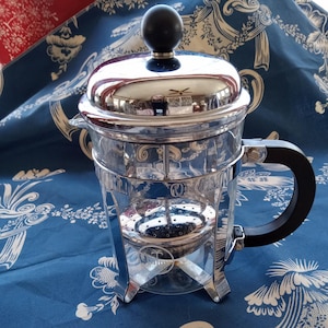 May include: A clear glass French press coffee maker with a shiny chrome lid, frame, and base. It has a black handle and a black knob on top. The coffee maker sits on a blue and white patterned surface.