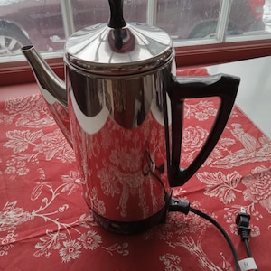 May include: A shiny, stainless steel electric coffee percolator with a black handle and lid knob. The coffee pot sits on a red tablecloth with a white floral pattern. A black power cord with a two-prong plug extends from the base.