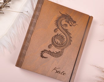 May include: A wooden journal with a dragon design engraved on the front. The journal is brown and has a black elastic closure. The name "Kate" is engraved on the bottom of the journal.