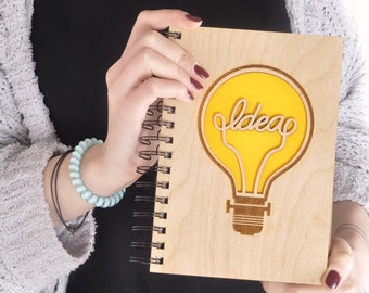 May include: A wooden spiral bound notebook with a light brown wood cover. The cover has a yellow lightbulb cutout with the word "idea" written inside.
