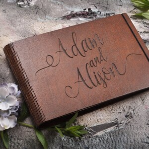 以下が含まれることがあります： 「AdamとAlison」と刻まれた茶色の木製のゲストブック。本は閉じており、グレーのざらざらした表面の上に置かれています。