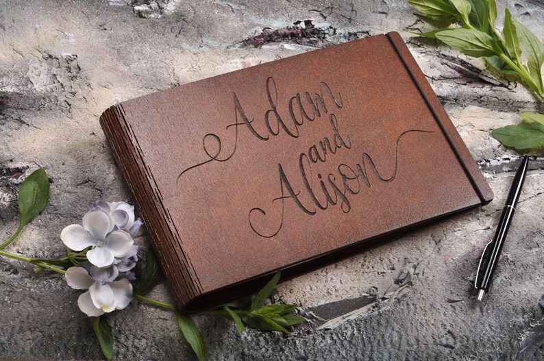 以下が含まれることがあります： 「AdamとAlison」と刻まれた茶色の木製のゲストブック。本は閉じており、グレーのざらざらした表面の上に置かれています。