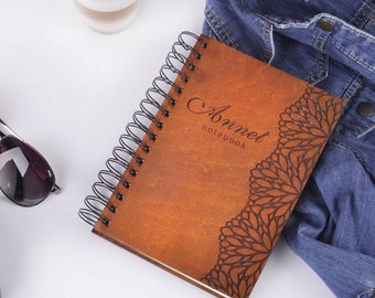 May include: A brown leather notebook with a black spiral binding. The notebook has a decorative floral design and the name "Anneet notebook" is engraved on the front cover.