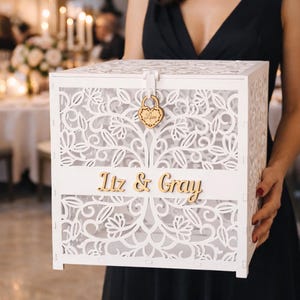 Könnte beinhalten: Eine weiße Holzkartenbox mit aufwendigen floralen Ausschnitten. Die Box hat ein herzförmiges Schloss mit dem Wort "Love" und den Namen "Liz & Gray" in goldfarbenen Buchstaben. Die Box wird von einer Person in einem schwarzen Kleid gehalten.