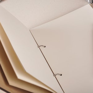May include: An open, brown leather bound journal with blank white pages. A silver pen with a black carbon fiber design is lying on the open pages.