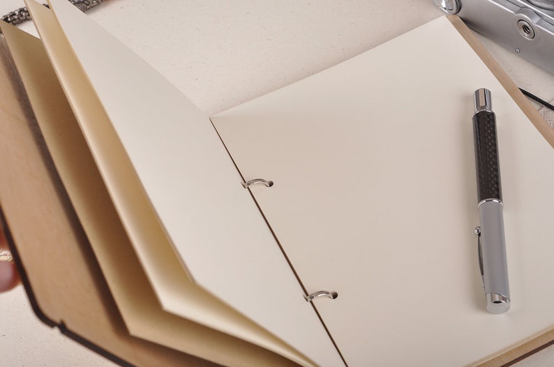 May include: An open, brown leather bound journal with blank white pages. A silver pen with a black carbon fiber design is lying on the open pages.