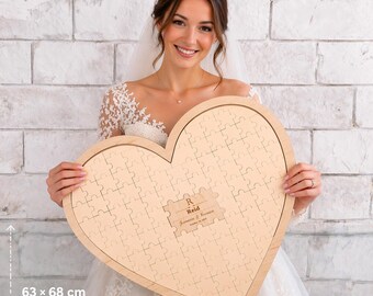 May include: A large, heart-shaped wooden puzzle, measuring 63 x 68 cm, with interlocking pieces. The puzzle has a light wood finish and includes a small rectangular section with the name "Reid" and a wedding date. A bride holds the puzzle.
