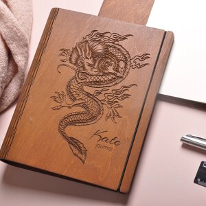 May include: A wooden journal with a dragon design engraved on the cover. The journal is brown and has the name "Kate Journal" engraved on the bottom.