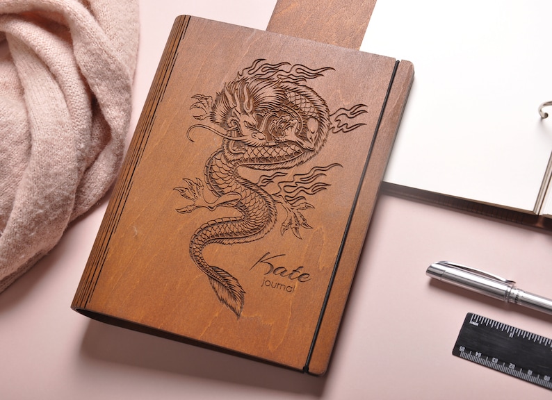 May include: A wooden journal with a dragon design engraved on the cover. The journal is brown and has the name "Kate Journal" engraved on the bottom.