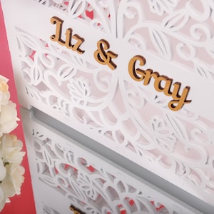 以下が含まれることがあります： 白いレーザーカットの装飾パネル、フローラルデザイン。 「Liz & Gray」の文字は金色の木製文字です。 白いバラの花束が左下にあります。 背景はピンク色です。