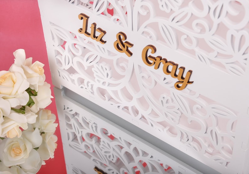 以下が含まれることがあります： 白いレーザーカットの装飾パネル、フローラルデザイン。 「Liz & Gray」の文字は金色の木製文字です。 白いバラの花束が左下にあります。 背景はピンク色です。