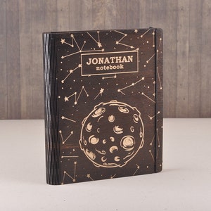 Può includere: Un taccuino in legno marrone scuro con un design a costellazione e un grande grafico della luna. Il taccuino ha il testo "JONATHAN notebook" inciso sul davanti.