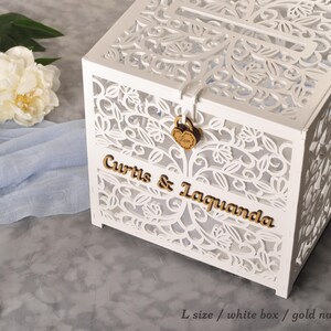 以下が含まれることがあります： 白いレーザーカットの木製カードボックス。ボックスには「Curtis & Laquanda」と書かれた金色の文字とハート型のロックが付いています。背景にはシャクヤクとキャンドルホルダーがあります。