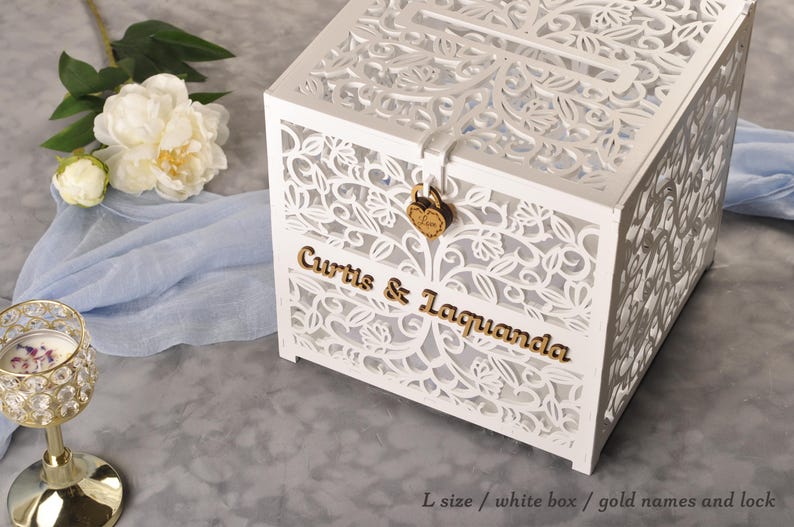 以下が含まれることがあります： 白いレーザーカットの木製カードボックス。ボックスには「Curtis & Laquanda」と書かれた金色の文字とハート型のロックが付いています。背景にはシャクヤクとキャンドルホルダーがあります。