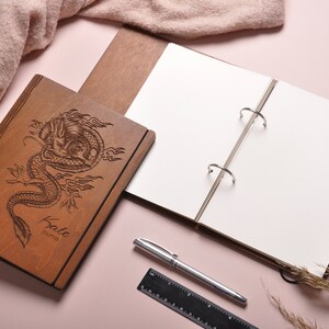 May include: A wooden journal with a dragon design engraved on the cover. The journal is open to reveal blank pages and a ring binder. The journal is personalized with the name "Katie" and the word "Journal".