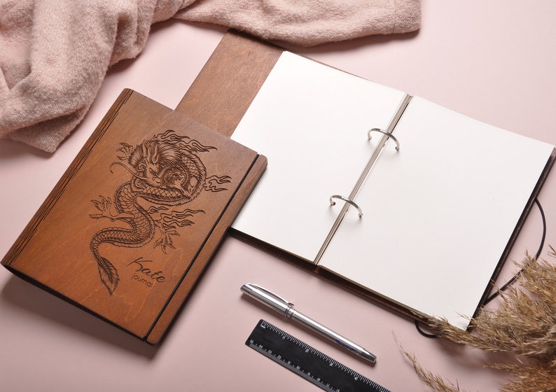 May include: A wooden journal with a dragon design engraved on the cover. The journal is open to reveal blank pages and a ring binder. The journal is personalized with the name "Katie" and the word "Journal".