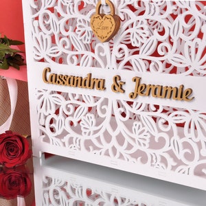 以下が含まれることがあります： 白い木製のカードボックスで、複雑な花の切り抜きがあります。「Cassandra & Jeramie」という名前が木製の文字で表示されています。「Love」という言葉が入った木製のハート型の南京錠が付いています。赤いバラとギフトバッグが背景にあります。