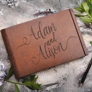 以下が含まれることがあります： 茶色の革張りゲストブックに「Adam and Alison」という名前が刻まれています。本は灰色のざらざらした表面に置かれ、ペンと緑の葉が置かれています。